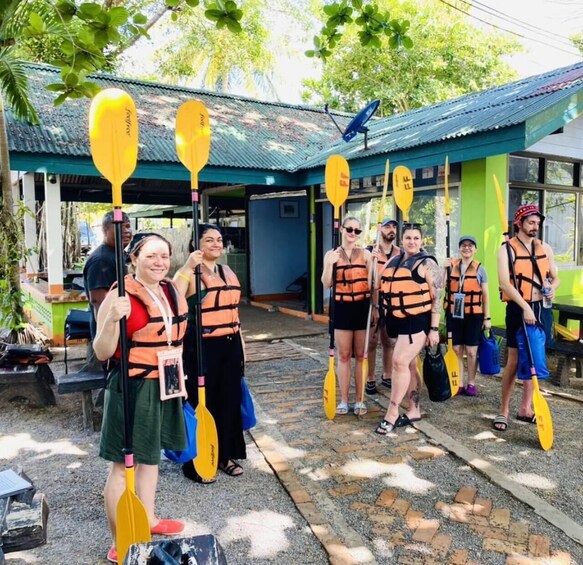 A group of people in life jackets holding paddles, standing outdoors near a building with a corrugated metal roof. 5