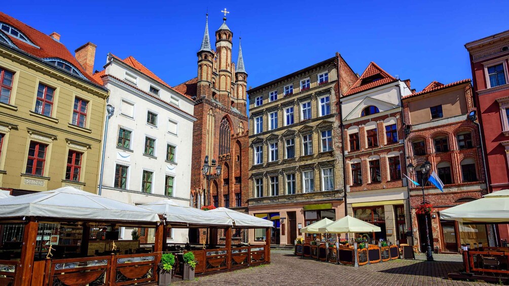 A cobblestone street with historic buildings, a church with twin spires, and outdoor seating areas with umbrellas. 1