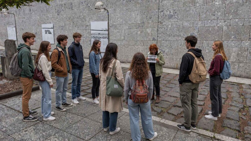 A group of people standing in a line on a stone-paved area with informational signs in the background. 3