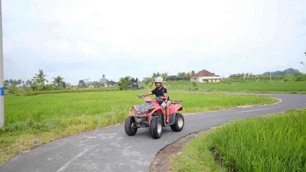 A person riding a red ATV on a rural road surrounded by green fields and a few houses. 5