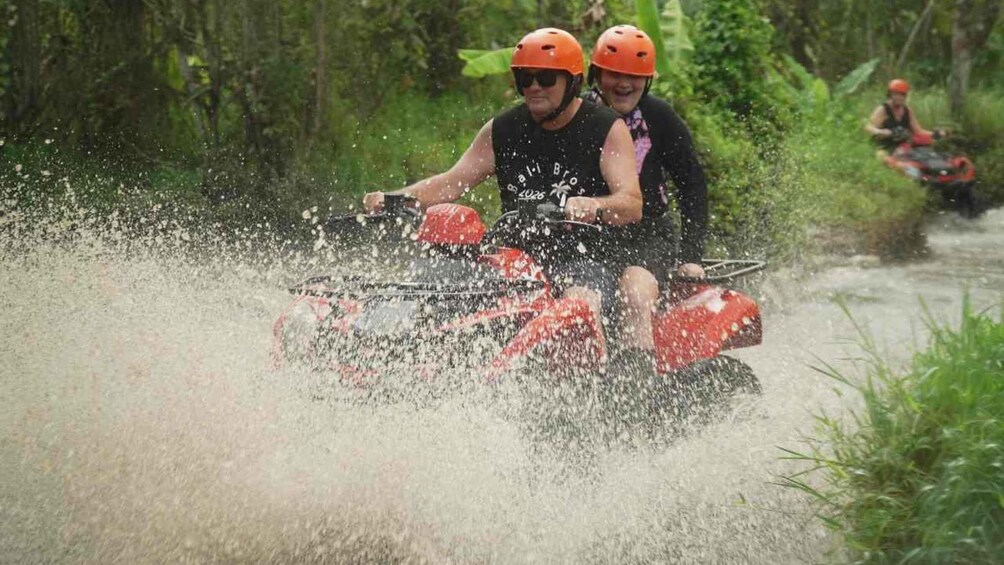 Two people on a red ATV in a forested area, splashing water. 1