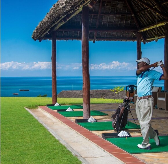A golf course with a view of the ocean, a thatched-roof pavilion, and a golfer preparing to swing. 1