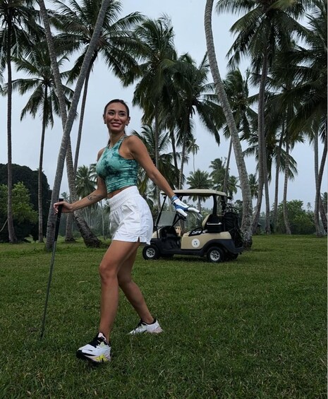 A person in a green top and white shorts walking on grass with a golf cart in the background. 5