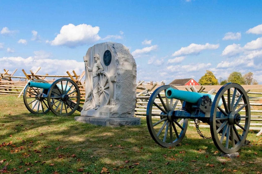 A cannon and a statue in a grassy field with a wooden fence and a red barn in the background. 1
