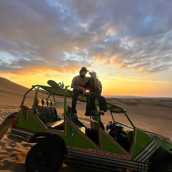 A green off-road vehicle with two people sitting on the roof, parked on a desert with sand dunes in the background. 5