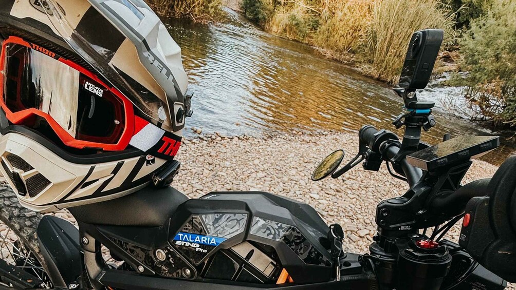 A motorcycle with a helmet and camera mounted on the handlebars, parked by a river. 2