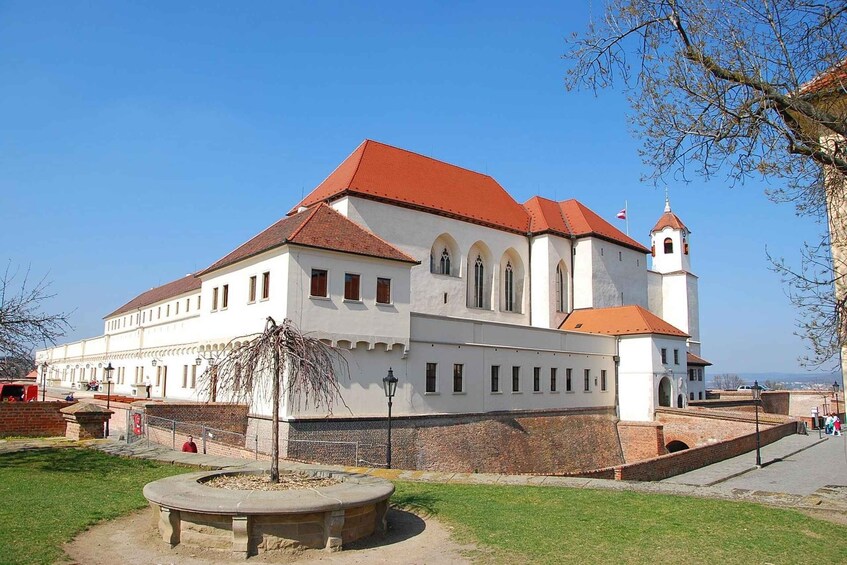 A large white building with a red roof and a tower, surrounded by a brick wall and a grassy area with a bench. 5