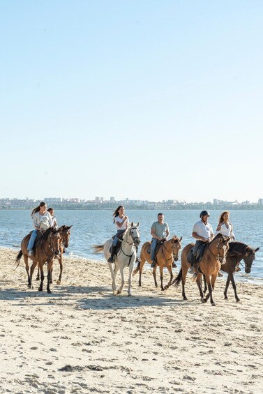 A group of people riding horses on a sandy beach with a body of water and a city skyline in the background. 4