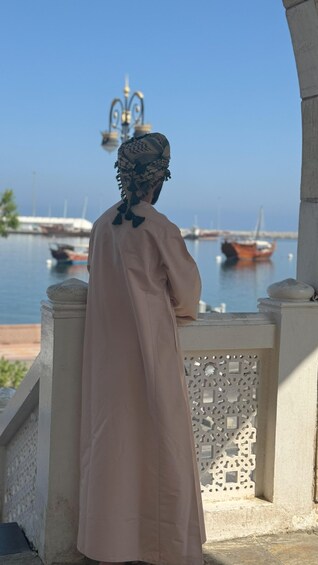 A person in traditional attire standing by a waterfront with boats and a decorative lamp in the background. 1