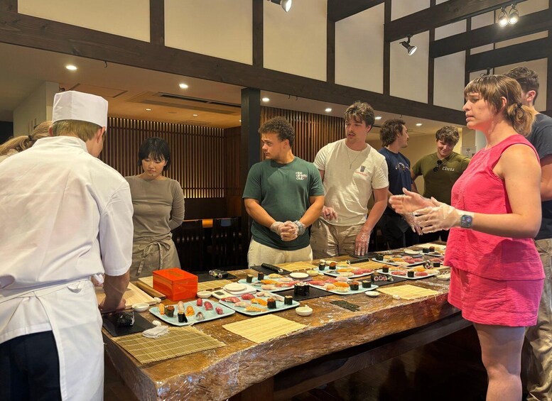 A group of people around a sushi bar with various dishes displayed. 3
