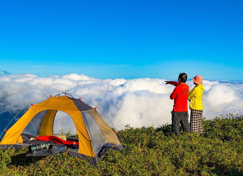 Two people standing by a yellow tent on a grassy hill, overlooking a sea of clouds. 2