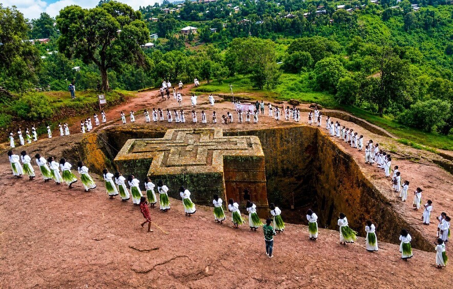 A group of people in white and green attire standing in a large, open-air stone structure with a cross design, surrounded by lush greenery and hills. 3
