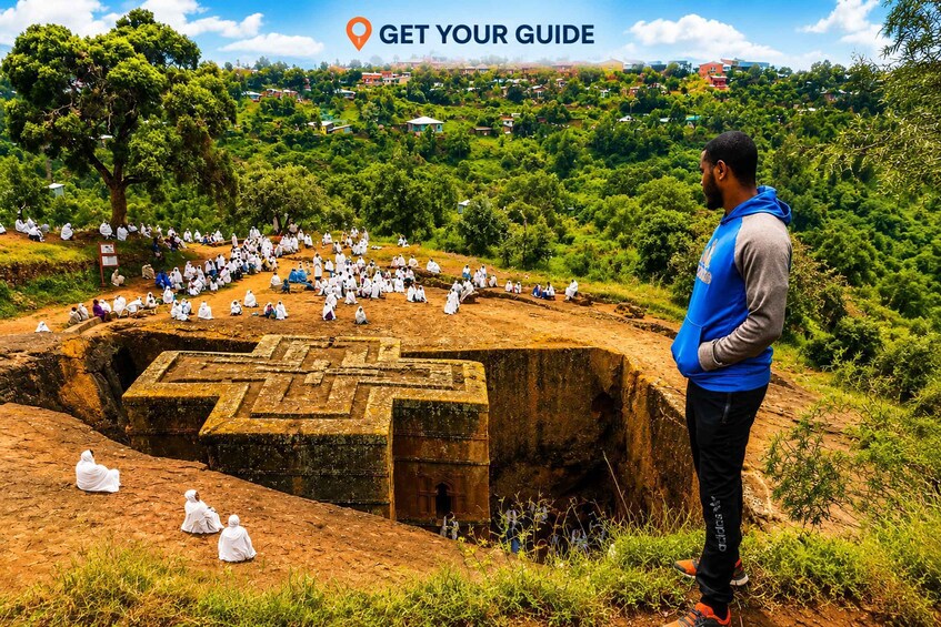 A large group of people in white robes gathered around a stone structure with a cross-like design, surrounded by lush greenery and a hilly landscape. 2