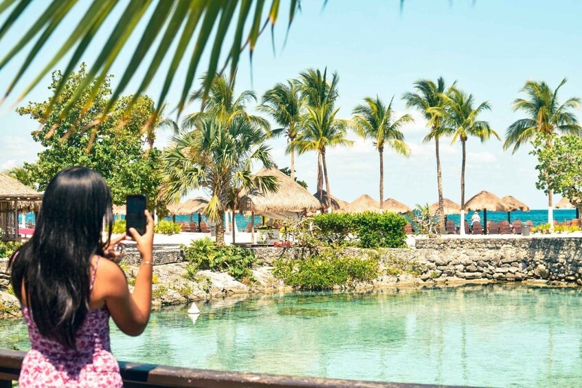 A woman taking a photo by a pool with palm trees and thatched-roof structures in the background. 4