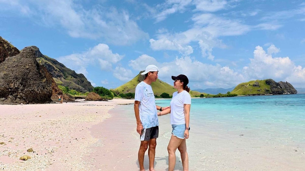 A couple on a beach with clear turquoise water, rocky outcrops, and green hills in the background. 2