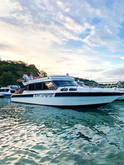 A white boat named 'Elo Voyage' docked at a marina with calm water and a forested shoreline in the background. 4