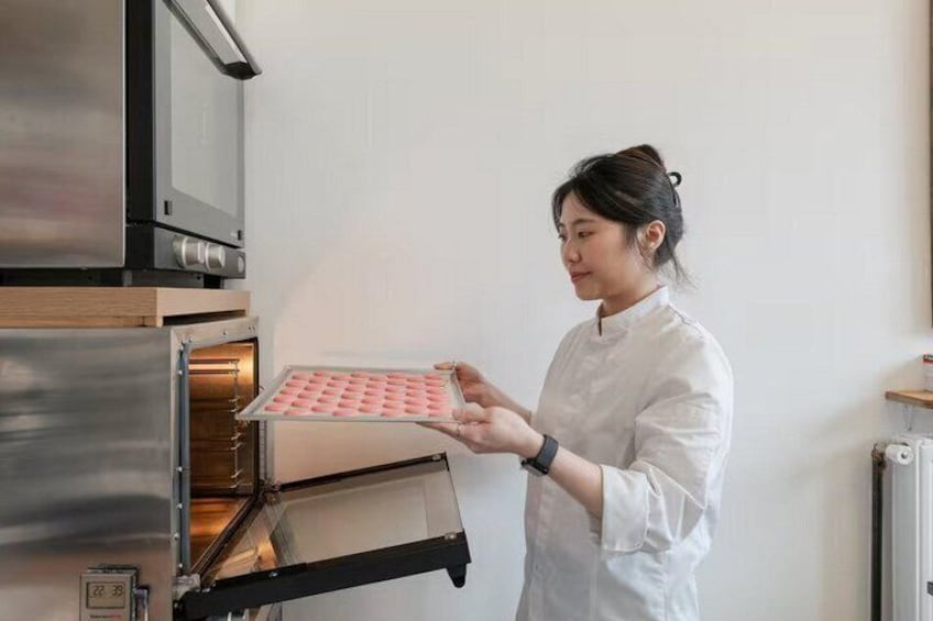 A person in a white chef's coat holding a tray with pink items, standing in front of a stainless steel oven. 1