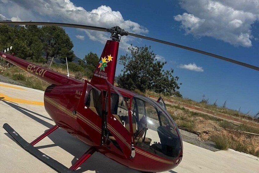 A red helicopter with 'EC-LO1' and a logo on its tail, parked on a paved surface with a clear sky and trees in the background. 1