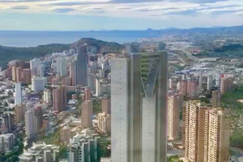 A cityscape with a prominent modern skyscraper, dense urban buildings, and a distant mountain range. 2