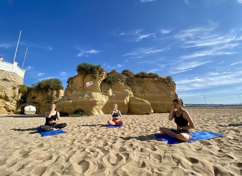 Three people meditating on blue mats on a sandy beach with a rocky cliff in the background. 3
