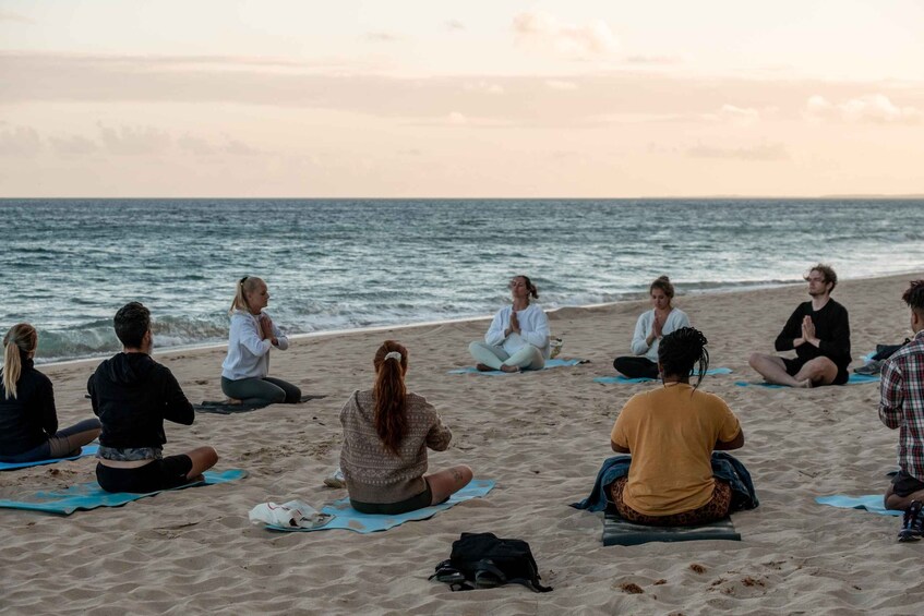 A group of people sitting on mats on a beach, facing the ocean. 2