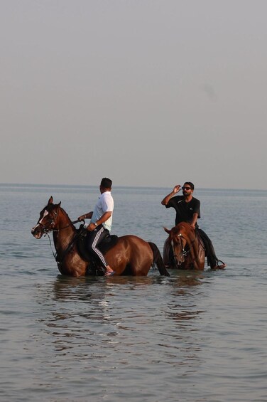 Two people riding horses in shallow water with a calm sea in the background. 3