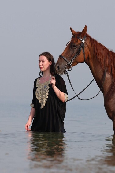 A person in black attire with gold embroidery leading a brown horse in shallow water. 1