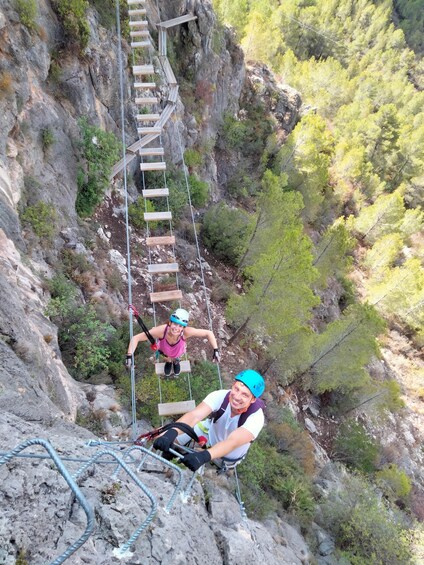 Two people on a wooden rope bridge in a mountainous area with trees. 3
