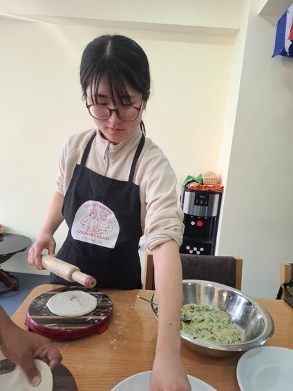 A person in a kitchen preparing food, wearing an apron with a logo, and using a rolling pin on dough. 3