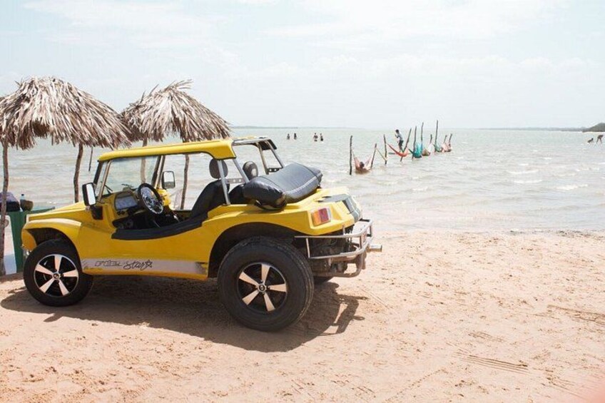 A yellow off-road vehicle parked on a sandy beach with palm trees and people in the water in the background. 2