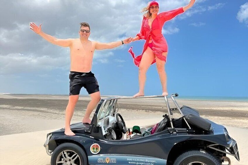 A man and a woman standing on a convertible car on a beach with a clear sky. 4