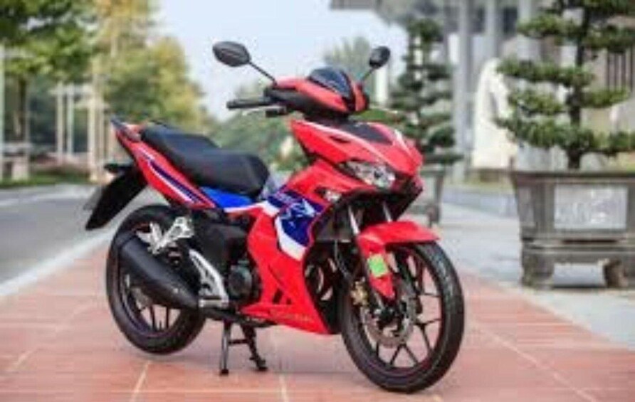 A red motorcycle parked on a paved surface with a blurred background of trees and a building. 1