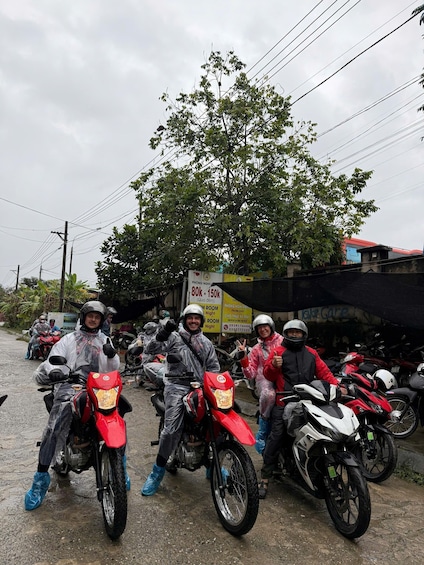 A group of motorcyclists on a wet road with parked motorcycles and a tree in the background. 3