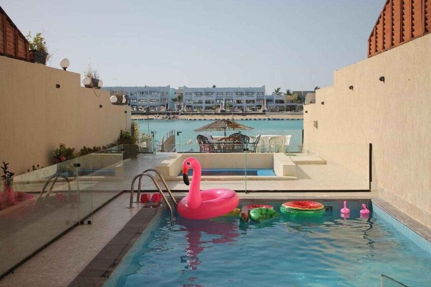 A swimming pool with a pink flamingo float, green and pink water toys, and a lounge area with a table and chairs. 2