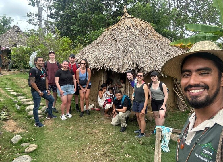 A group of people standing in front of a thatched-roof hut in a grassy area with trees in the background. 1