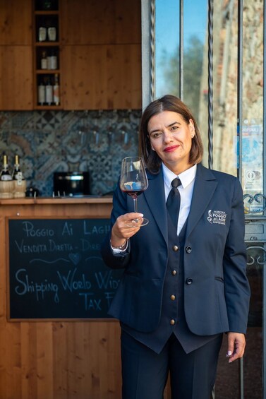 A person in a dark suit holding a wine glass, standing in front of a chalkboard with writing and a wooden counter with bottles. 1