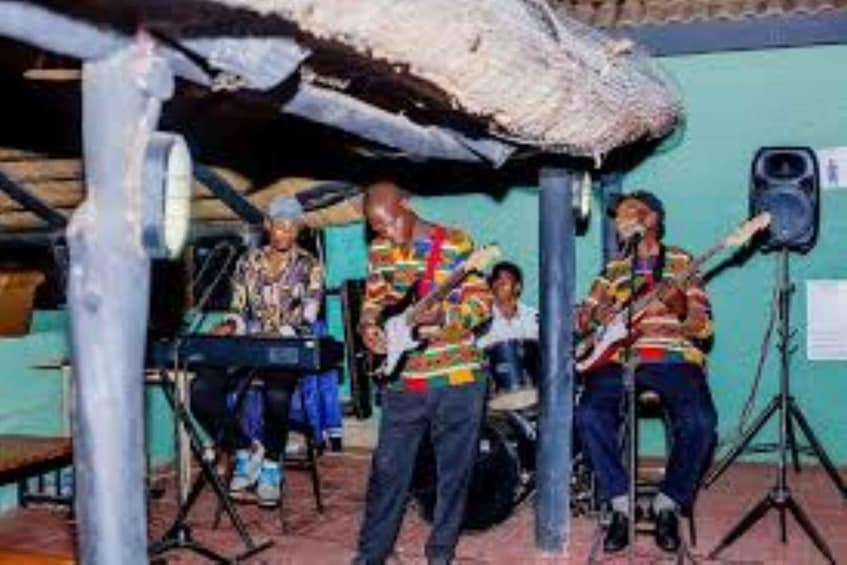 A group of musicians performing in an outdoor setting with a thatched roof and a microphone stand. 5