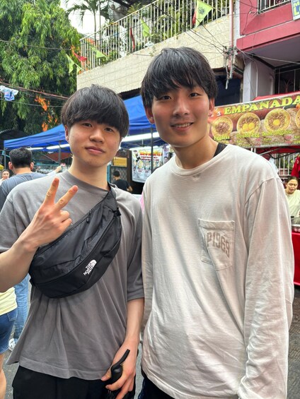 Two individuals standing in an outdoor market with a food stall in the background. 2