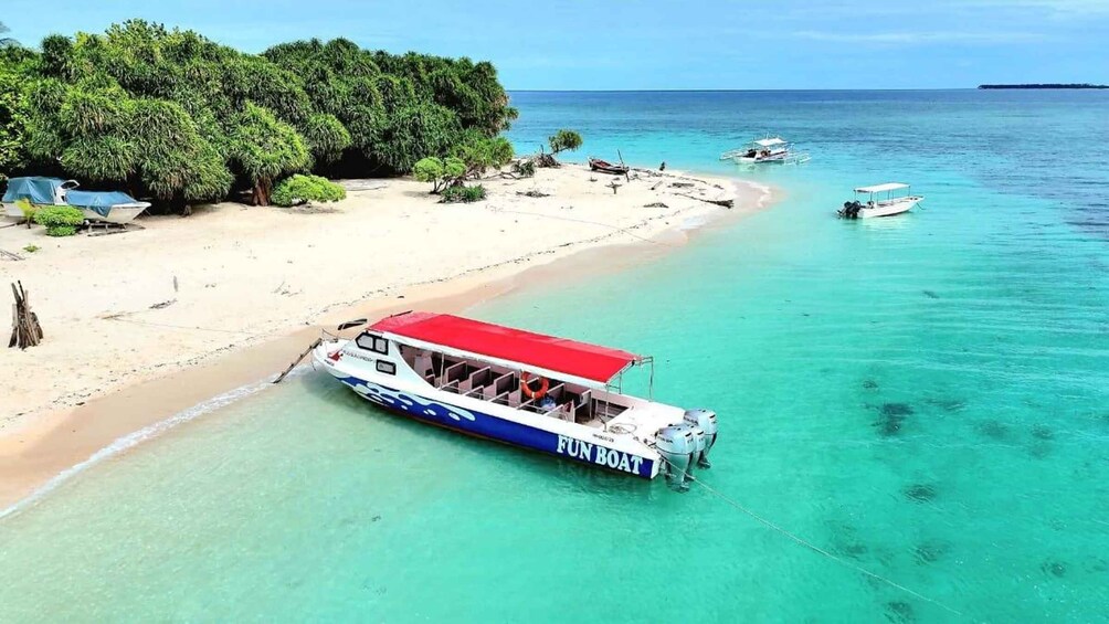 A beach with clear turquoise water, a boat named 'FUN BOAT' docked on the shore, and a forested area in the background. 1