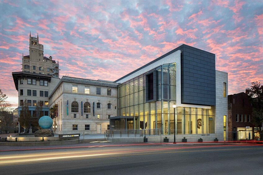 A modern building with a glass facade, a historic tower, and a large sculpture in front. 1
