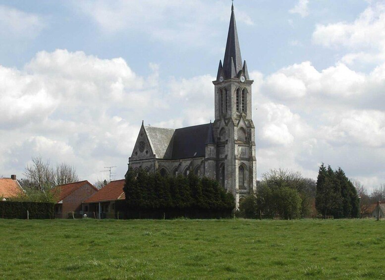 A church with a tall spire and a clock tower, surrounded by a green field and a few houses. 3