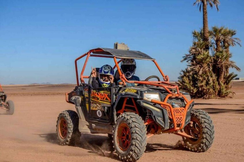 A desert off-road vehicle with two occupants, large tires, and a desert landscape with palm trees. 2