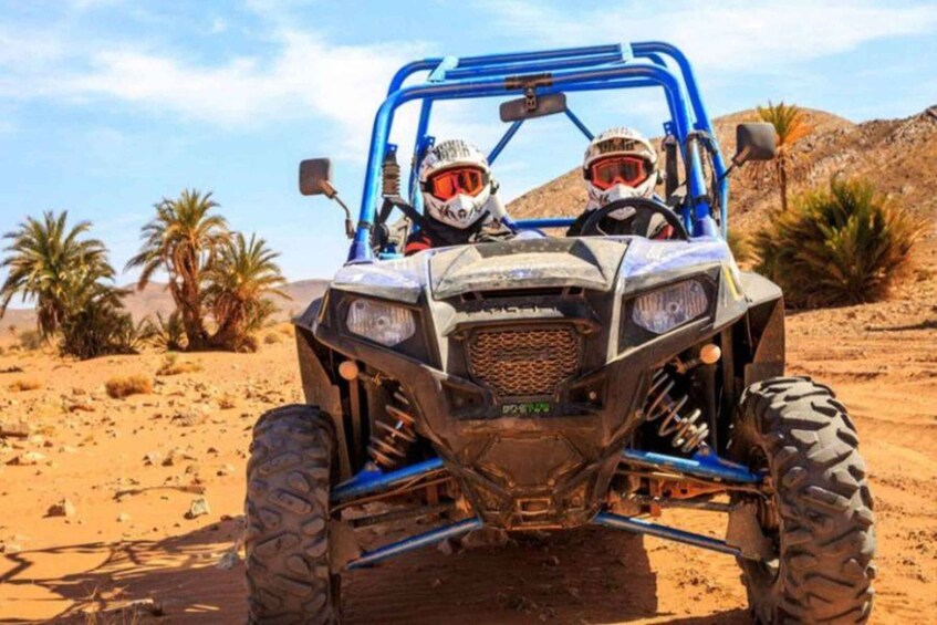 A blue off-road vehicle with two occupants in a desert landscape with sparse vegetation. 1