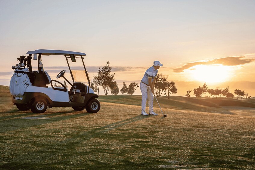 A golf course at sunset with a golf cart and a golfer preparing to take a shot. 1