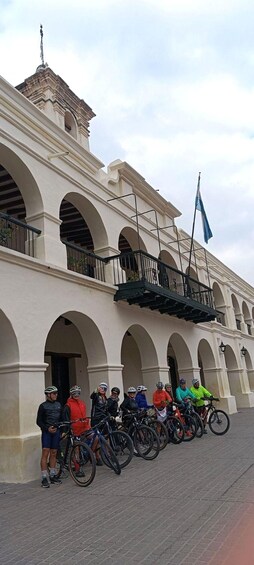 A group of cyclists standing in front of a white building with arches and a balcony. 2