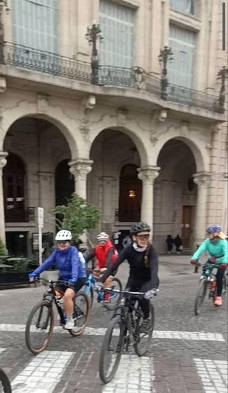Cyclists riding on a cobblestone street with a historic building featuring arches in the background. 3