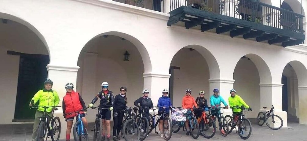 A group of cyclists standing in an outdoor area with arches and a balcony. 5