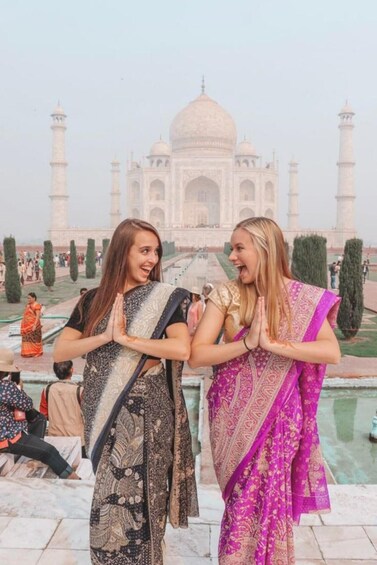 Two women in traditional Indian attire, one in a black and white saree and the other in a pink saree, standing in front of the Taj Mahal with their hands in a prayer position. 5