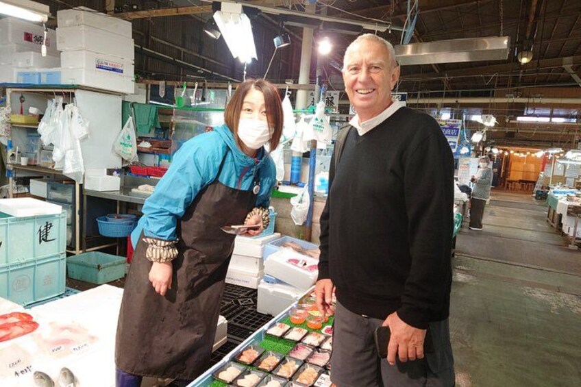 A person in a blue shirt and apron standing at a display with various food items, and another person in a black sweater and jeans in the background. 5