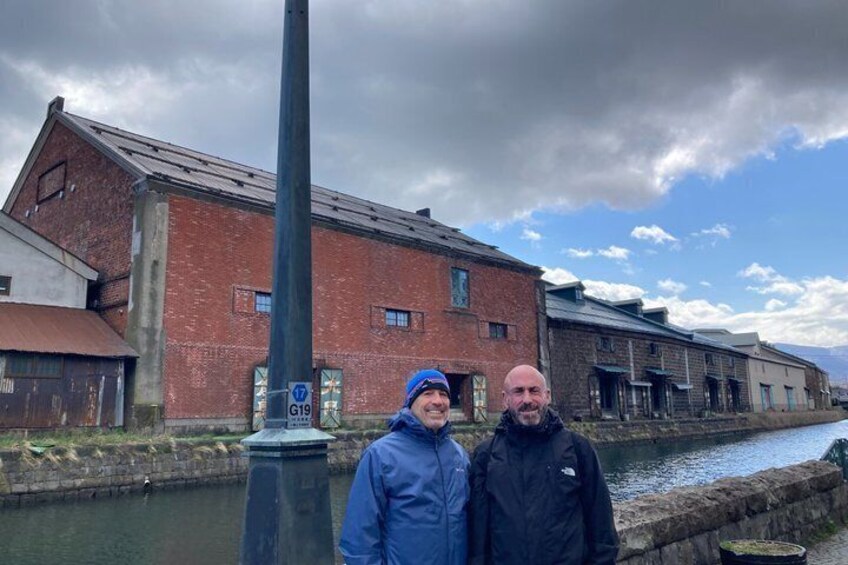 Two people standing in front of a brick building with a canal and a sign indicating a route. 4